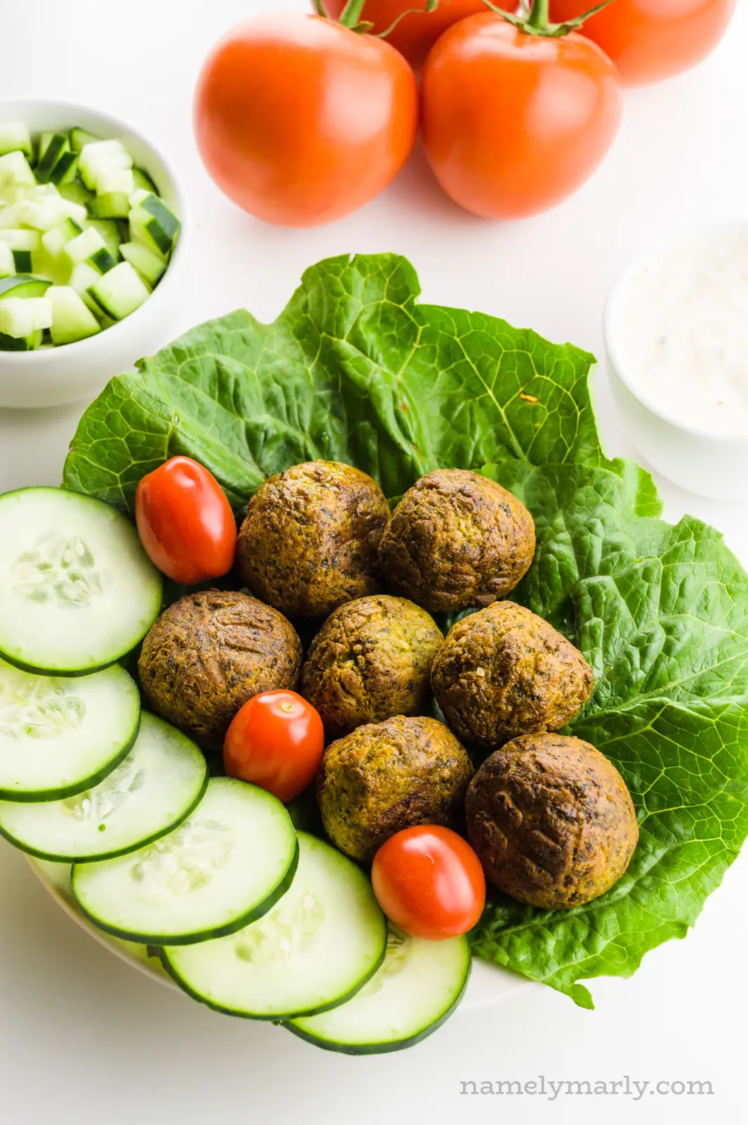 Air Fryer Falafels sit on a plate with veggies, such as lettuce and cucumbers. A bowl of sliced cucumbers is behind it along with a group of tomatoes.