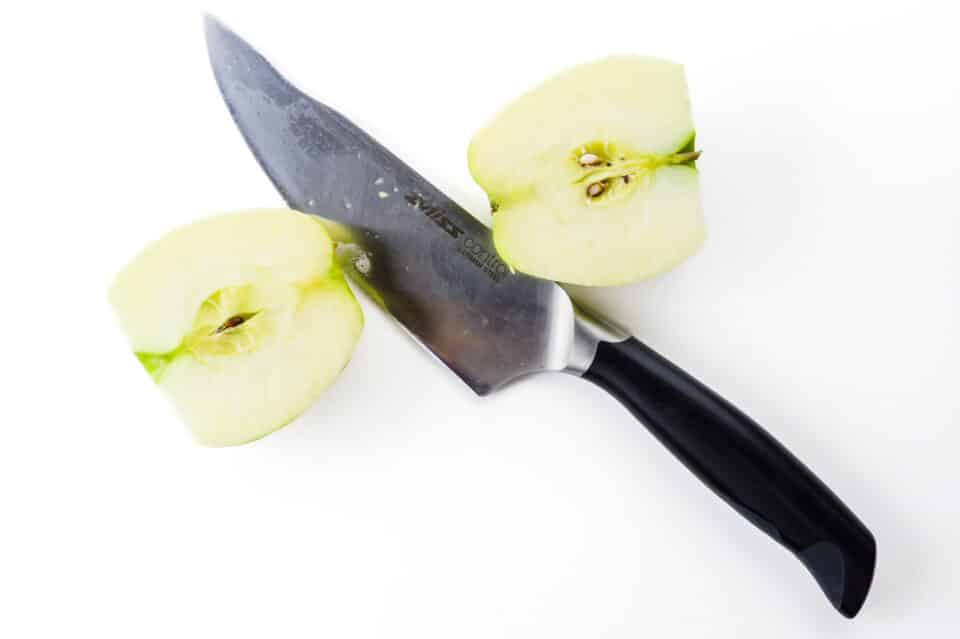 An apple has just been cut in half. The knife sits between the two sides of the cut apple. It's a step for cutting apples for apple pie.
