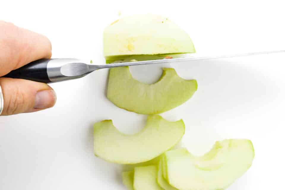 A hand holds a knife and is using it to slice apples into thin slices.