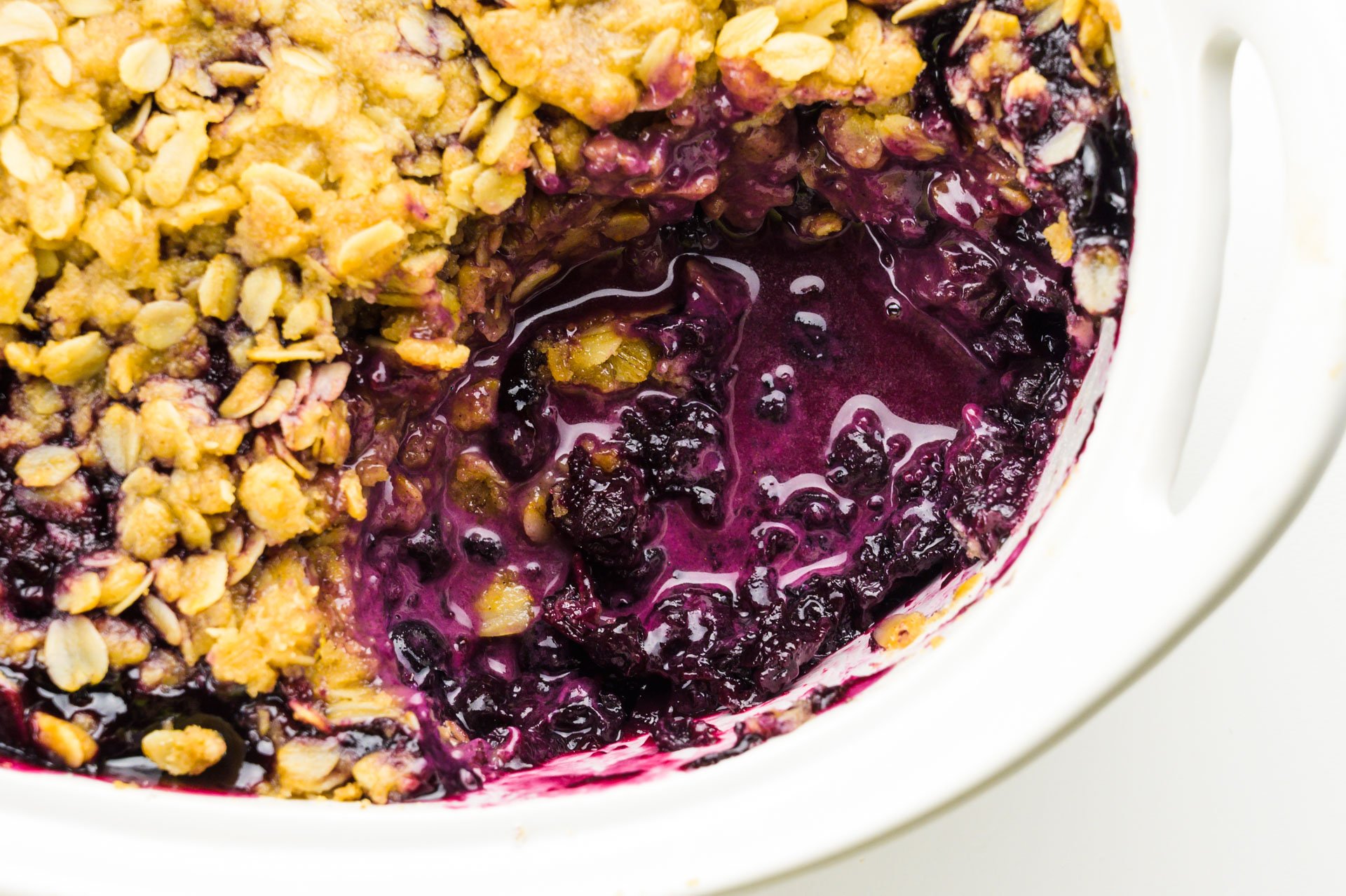 Looking down on a casserole dish full of blueberry crumble. A bite has been taken out, revealing, blueberry sauce at the bottom.
