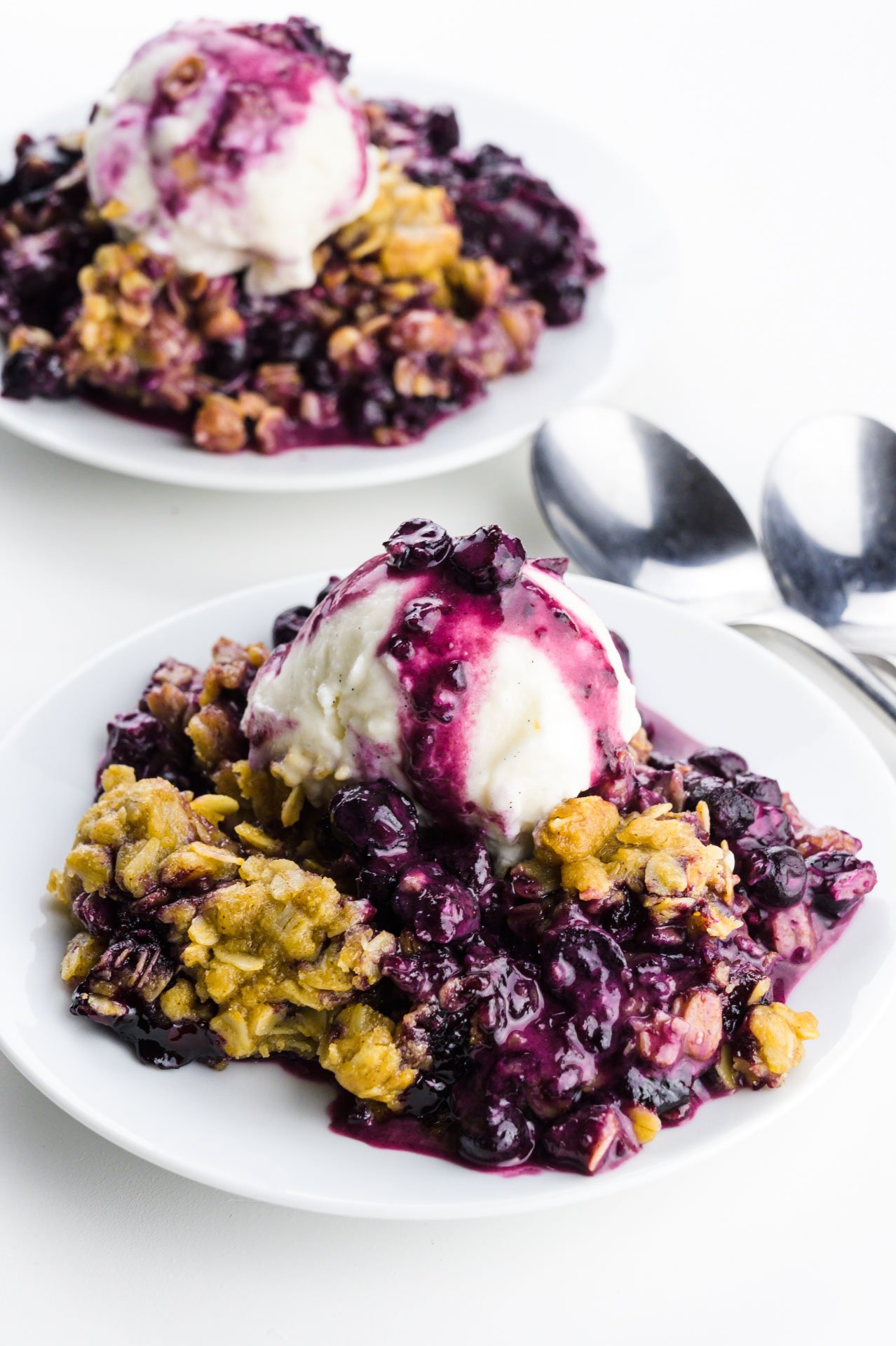 Two plates full of blueberry crumble with ice cream on top. There are two spoons between them.