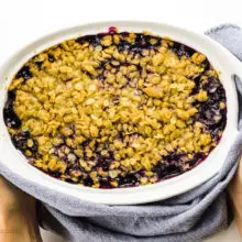 Hands hold a towel around a freshly baked batch of blueberry crumble.