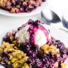 Two plates full of blueberry crumble with ice cream on top. There are two spoons between them.