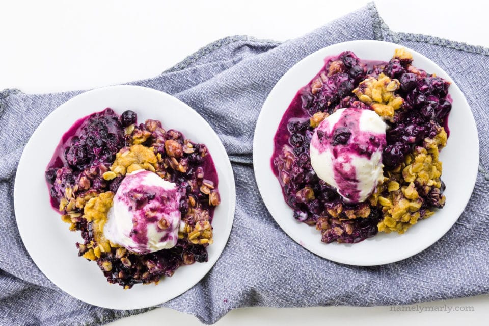 Two plates of blueberry crumble have ice cream on top and blueberry sauce drizzled on top. They are sitting on a kitchen towel.
