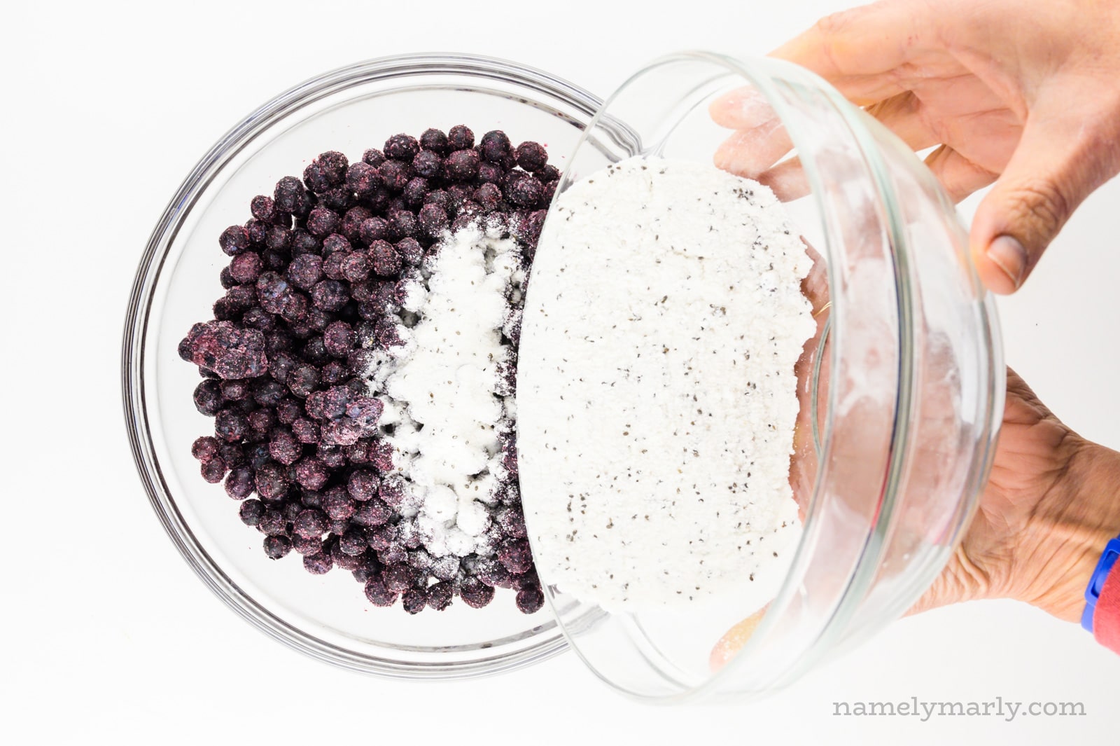 Two hands hold a bowl of flour mixture, pouring it in with a bowl of frozen blueberries.