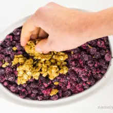 A hand holds crumble mixture and is spreading it across blueberries in a casserole dish.