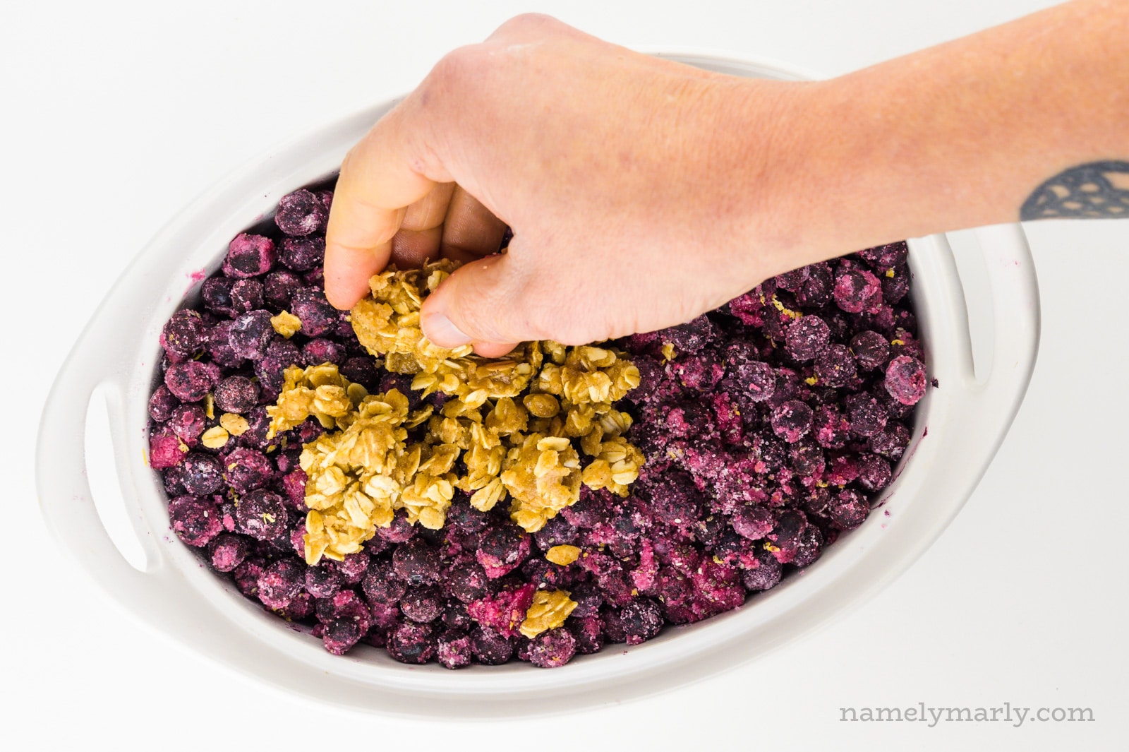 A hand holds crumble mixture and is spreading it across blueberries in a casserole dish.