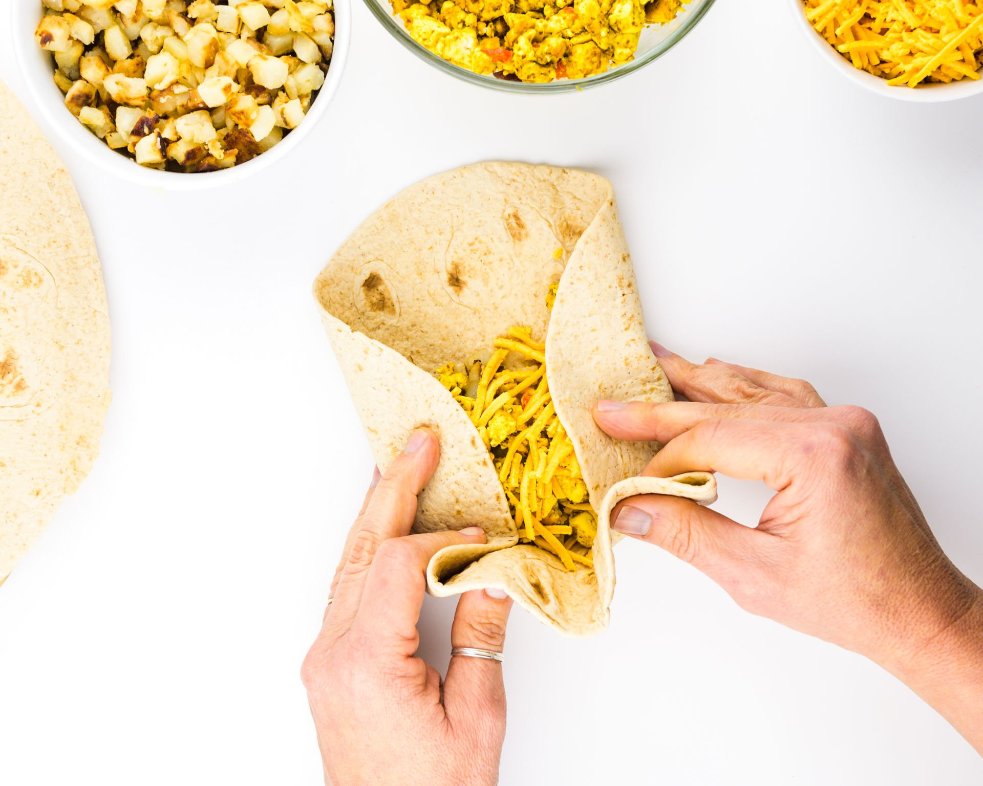 Two hands grasp a tortilla, rolling it into a breakfast burrito. There are other ingredients around the tortilla.