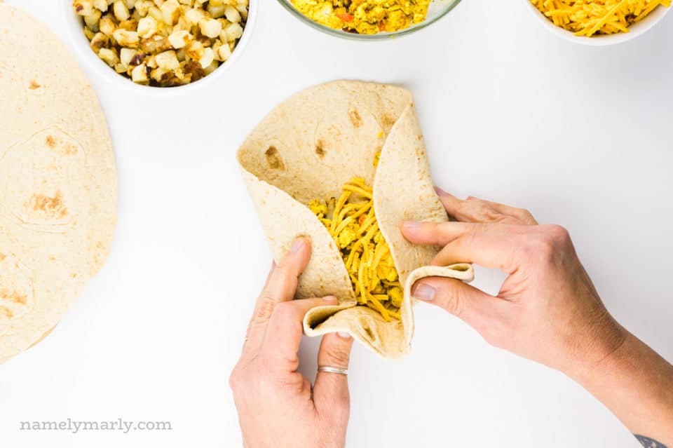 Two hands grasp a tortilla, rolling it into a breakfast burrito. There are other ingredients around the tortilla.