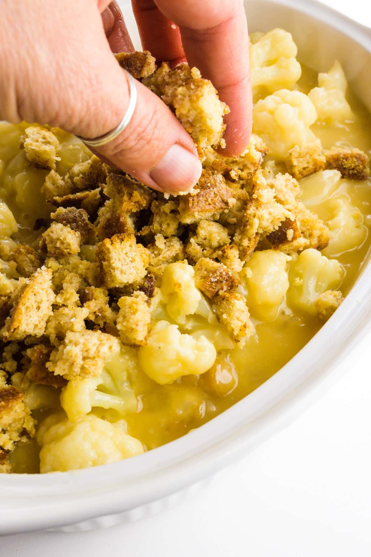 A hand holds bread crumbs and is dropping them over a cauliflower mixture in a casserole dish.