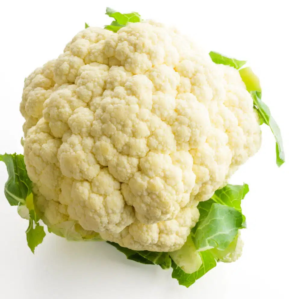 A head of cauliflower with green leaves intact sits on a white counter.
