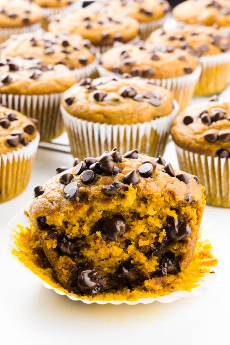 A pumpkin muffin is cut in half with lots of chocolate chips inside and on top. There are several muffins behind it too.