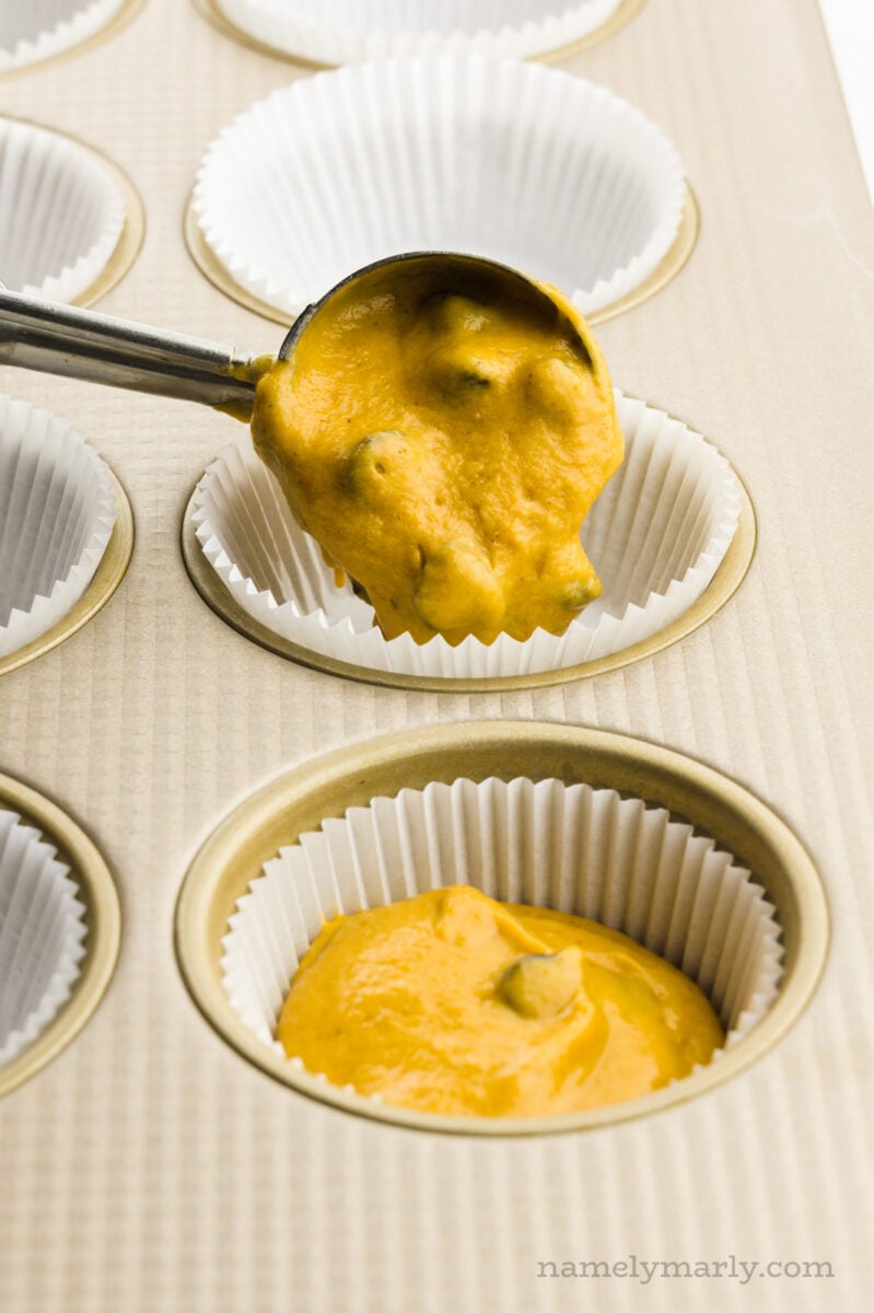 A cookie scooper is pouring batter into an muffin compartment lined with a paper.