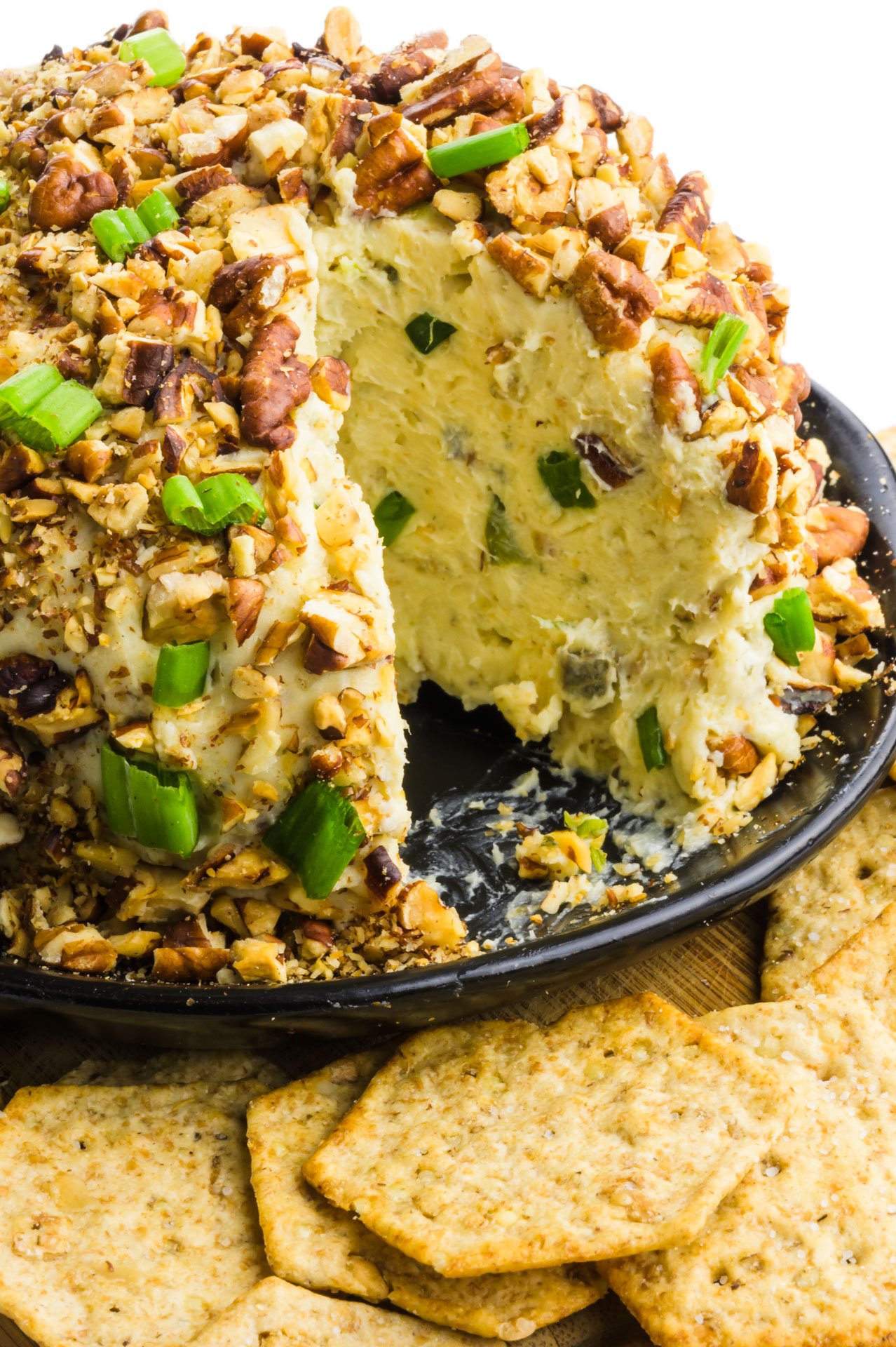 A cheeseball sits next to crackers and has a wedge cut out of it. 