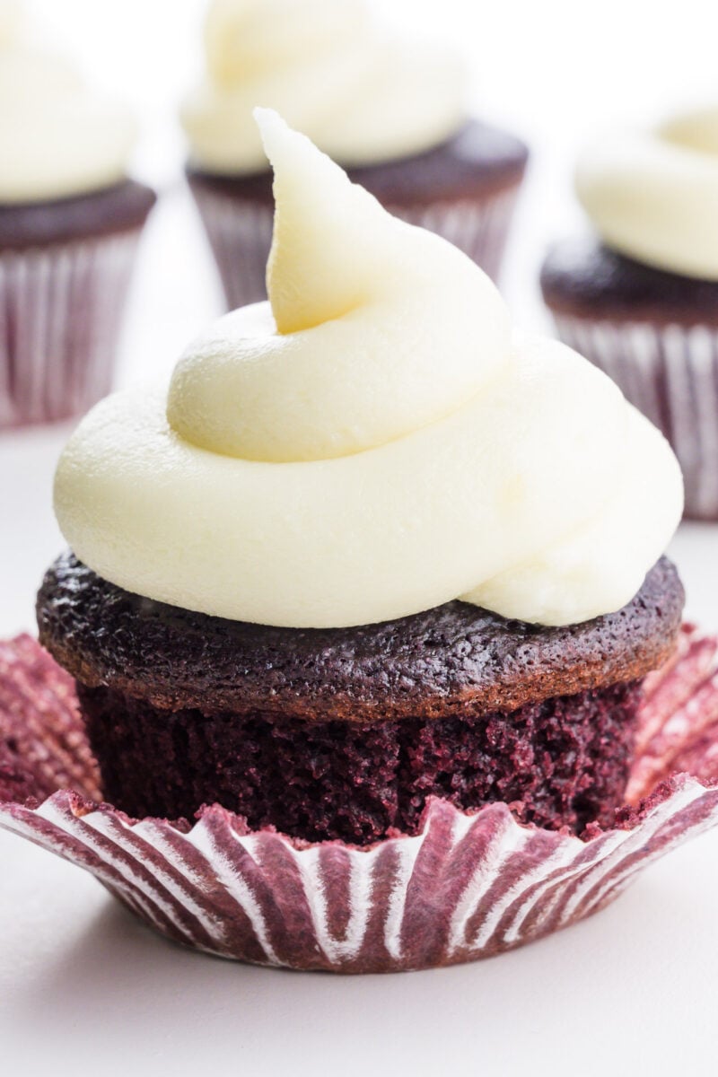 A red velvet cupcake is topped with vegan cream cheese frosting. There are more cupcakes behind it.