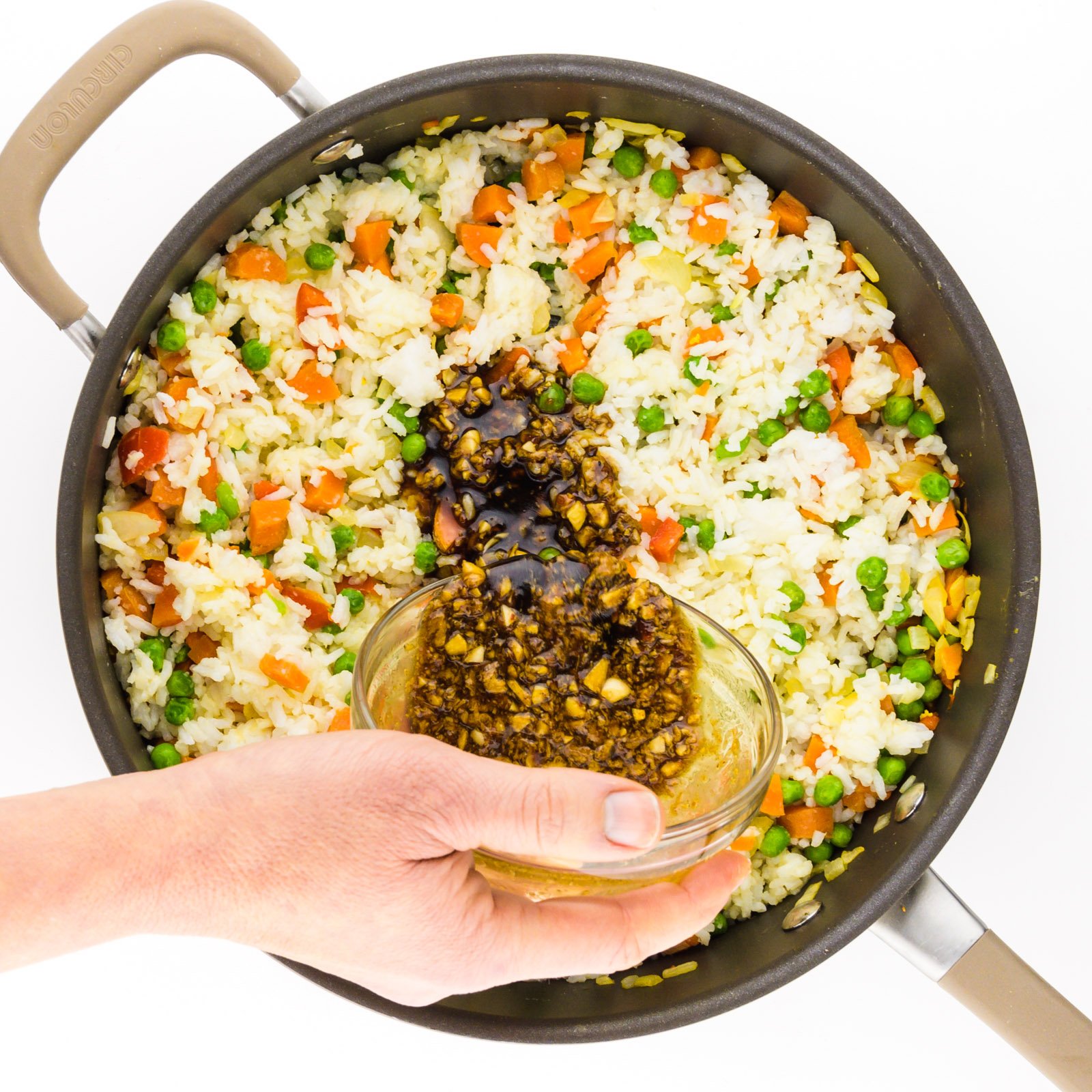A hand holds a bowl of sauce and is pouring it into a skillet full of rice and veggies.