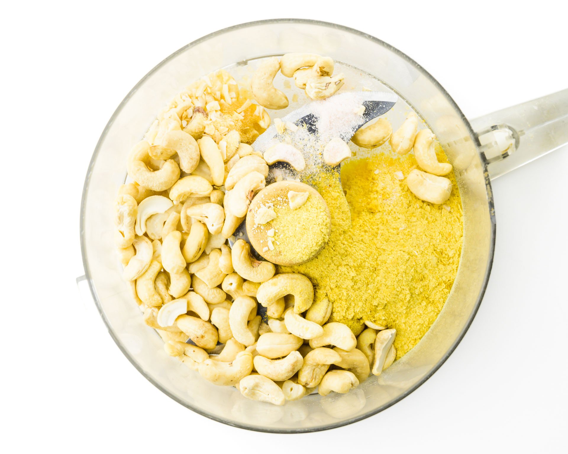 Cashews, nutritional yeast flakes, and spices are in the bowl of a food processor.