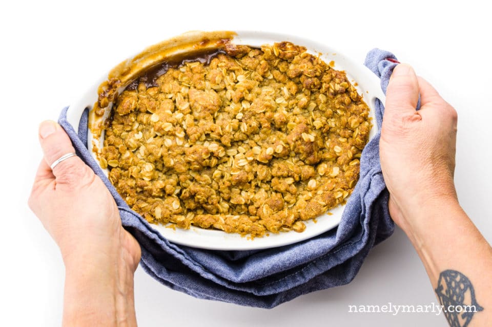 Two hands hold a blue kitchen towel at both ends of a casserole dish full of an apple dessert.
