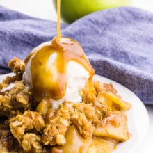 A plate holds vegan apple crisp with ice cream on sauce. Caramel sauce is being poured over the top. A blue napkin and green apple is behind it.