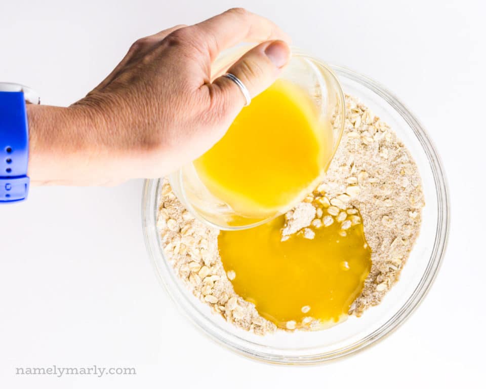 A hand holds a bowl of melted butter and is pouring it over a bowl full of a flour mixture.