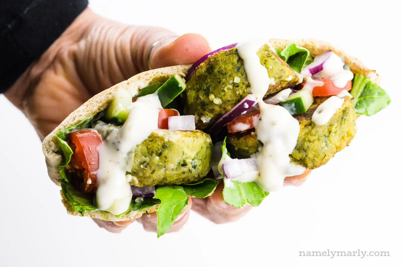 A hand holds a baked falafel sandwich with greens and sauce.
