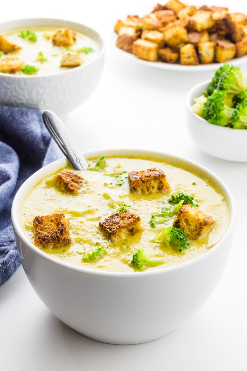 A bowl of broccoli soup with croutons on top. There's a bowl of broccoli behind it, another bowl of soup and more croutons.