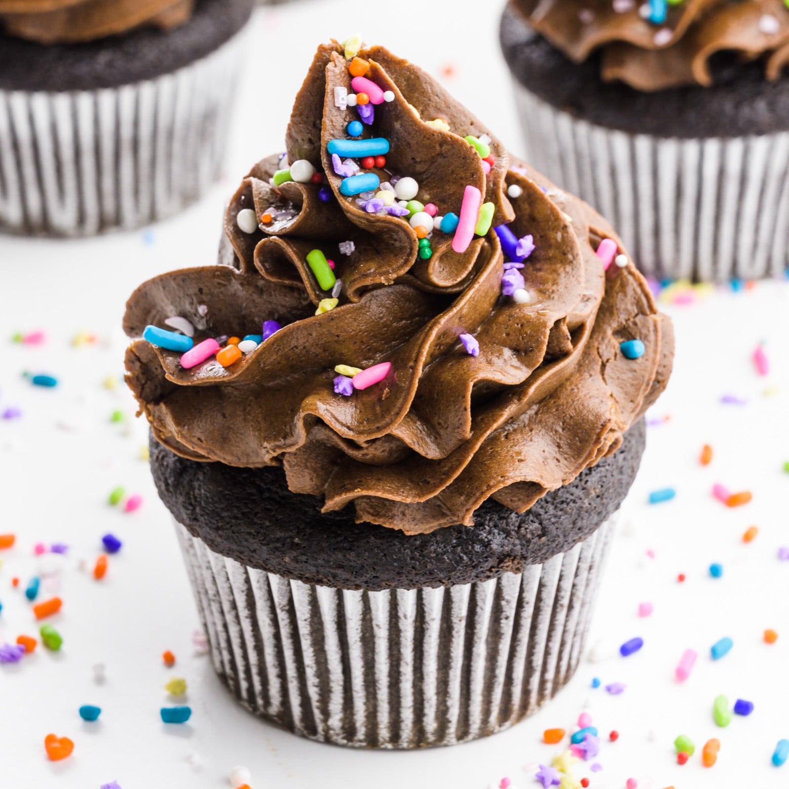 A chocolate cupcake has lots of colorful sprinkles on it and around it.