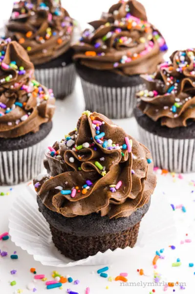 A group of several vegan chocolate cupcakes have colorful sprinkles on top.