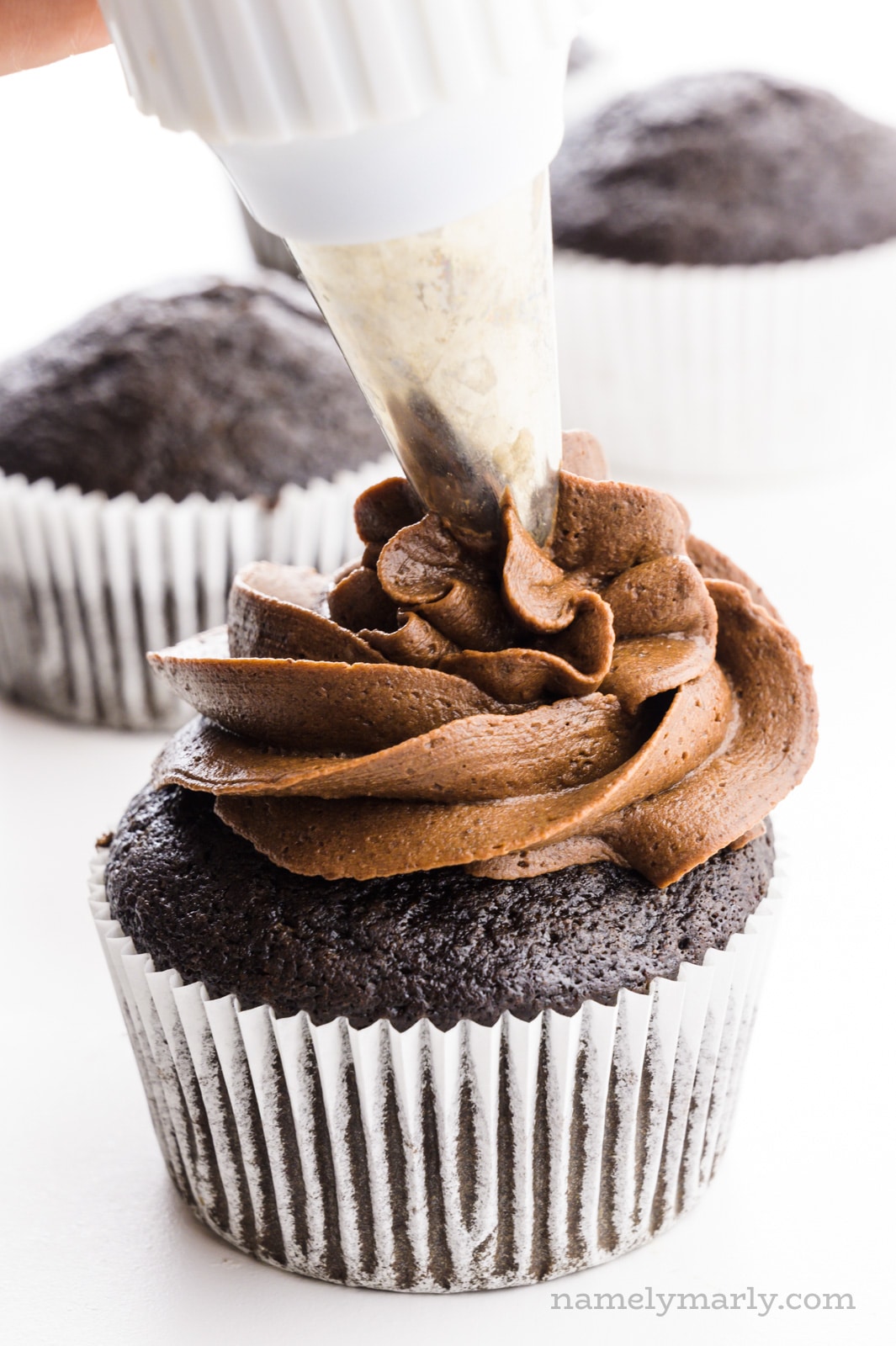 A hand holds a piping bag full of chocolate frosting and is distributing it over a chocolate cupcake.