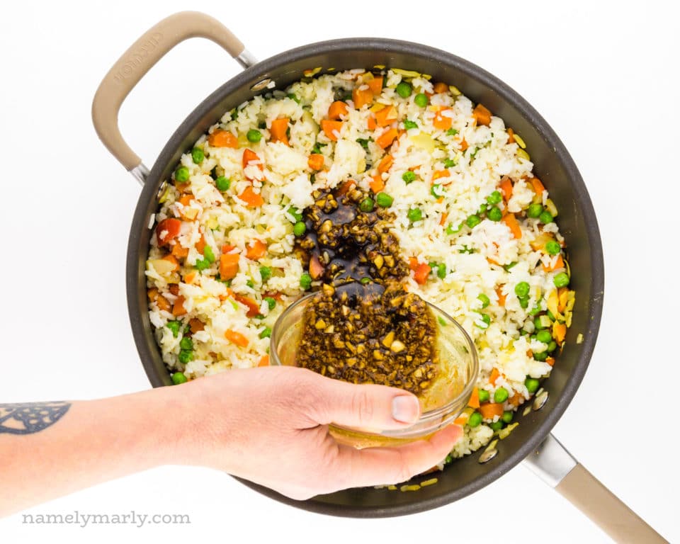 A hand holds a bowl of sauce and is pouring it into a skillet full of rice and veggies.