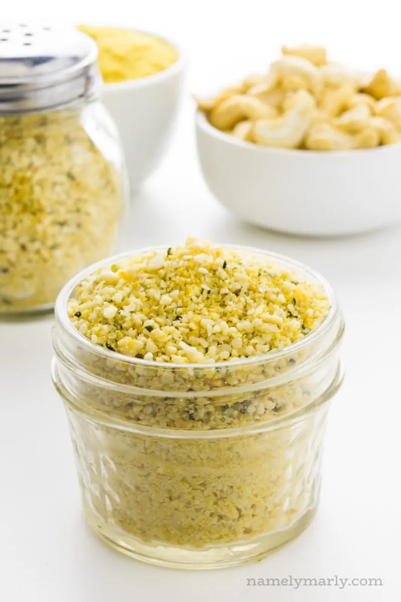 A glass jar full of vegan parmesan sits in front of a bowl of cashews, another bowl of nutritional yeast flakes, and a shaker full of more parmesan.