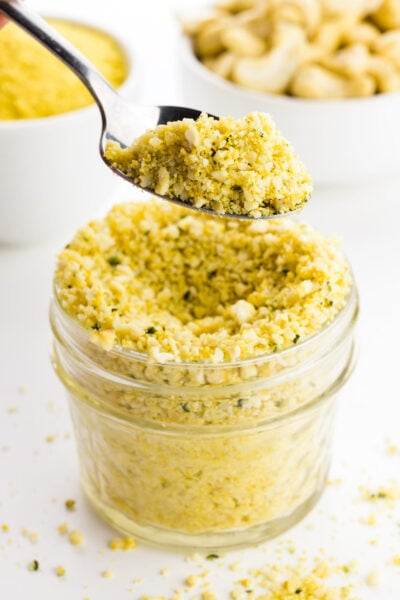 A hand holds a spoonful of cashew parmesan having over a glass jar full of it wit cashews behind it.