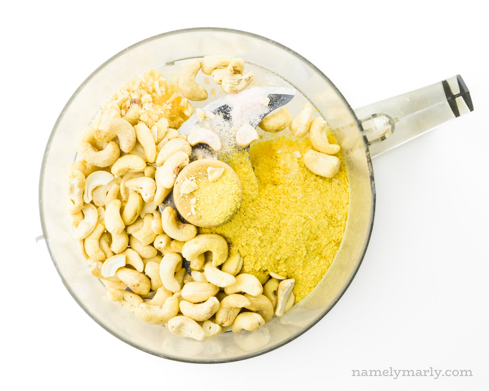 Cashews, nutritional yeast flakes, and spices are in the bowl of a food processor.