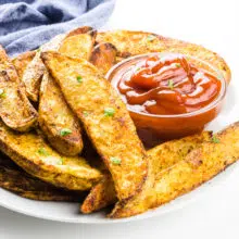 A plate holds several crispy air fryer potato wedges next to a bowl of ketchup.