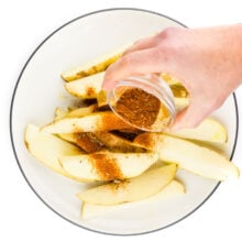 A hand holds a small bowl of spices, pouring it over potatoes wedges on a plate.