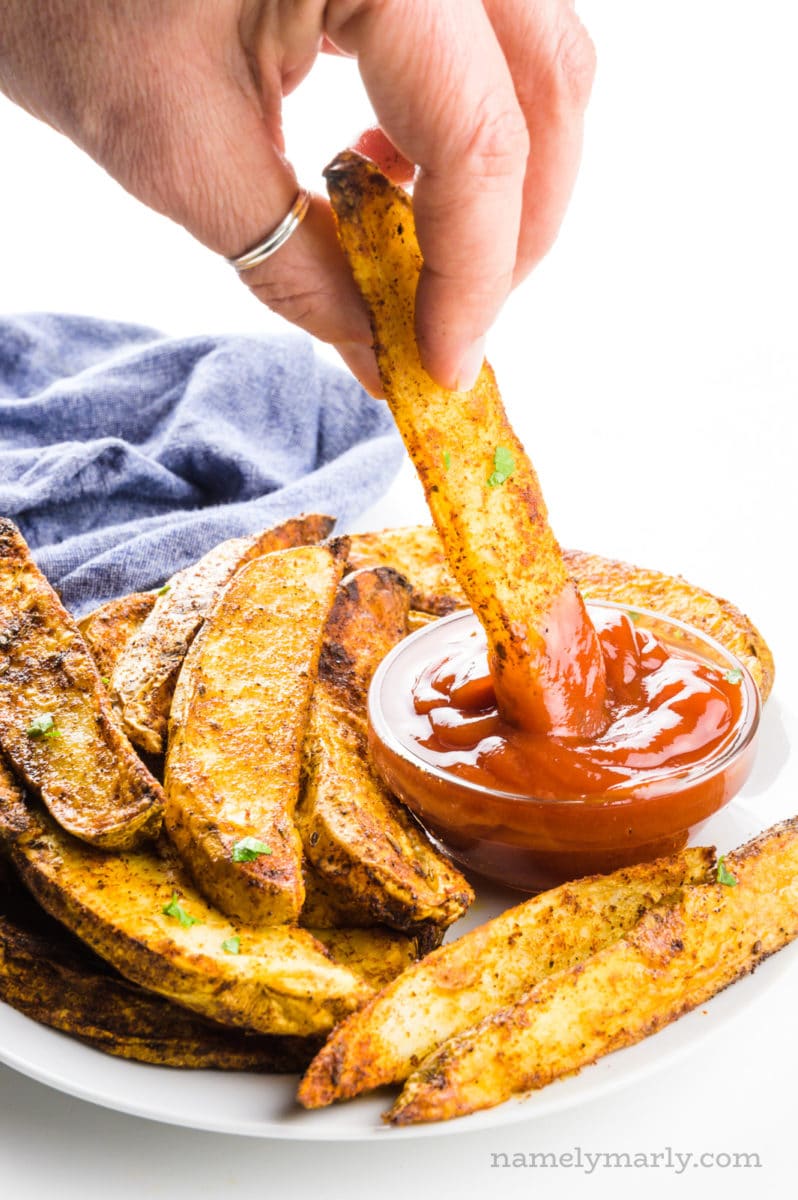 A hand holds a potato wedge dipping it in a bowl of ketchup on a plate with more fried potatoes.