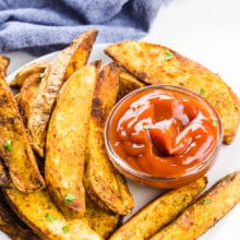 Air fryer potato wedges on a plate with a bowl of ketchup and a blue kitchen towel behind it.