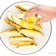 A hand holds a small bowl of oil, drizzling it over cut potatoes.