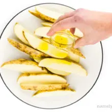 A hand holds a small bowl of oil, drizzling it over cut potatoes.