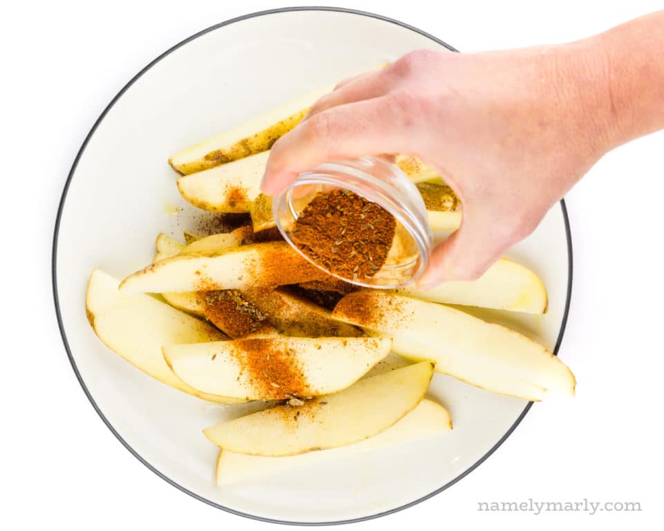 A hand holds a small bowl of spices, pouring it over potatoes wedges on a plate.