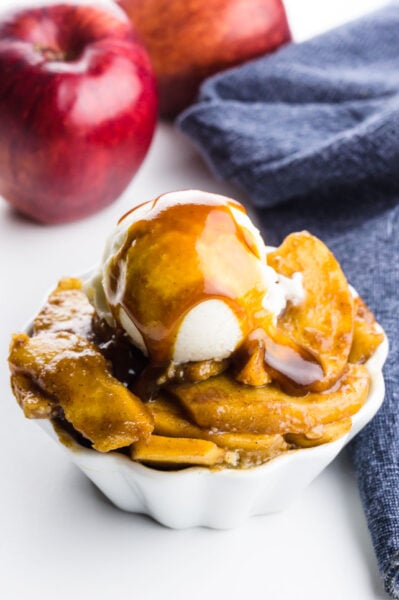 A bowl of baked apple slices is topped with ice cream and caramel sauce. There are apples behind it and a blue kitchen towel.