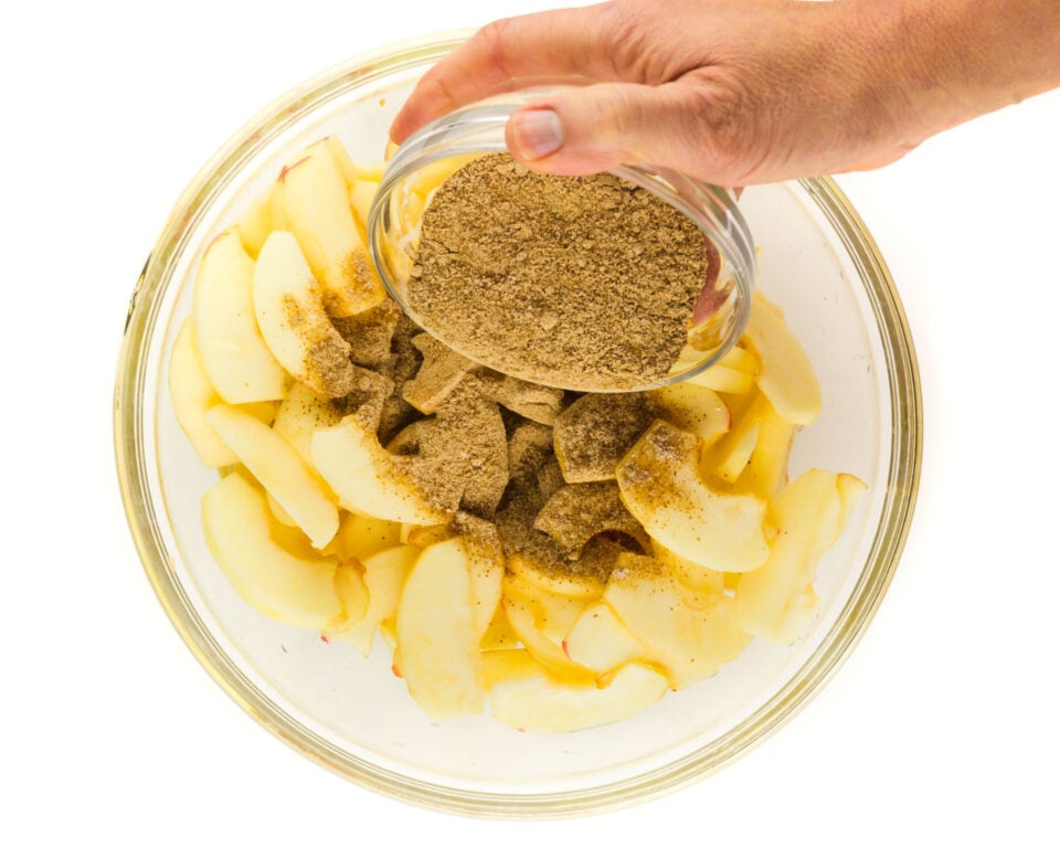 A hand pours a bowl of cinnamon spices over a bowl of freshly sliced apples.