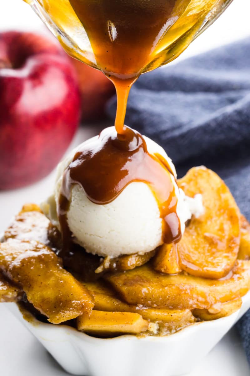 Caramel sauce is being drizzled over ice cream atop baked apple slices. There is an apple and a blue kitchen towel behind it.