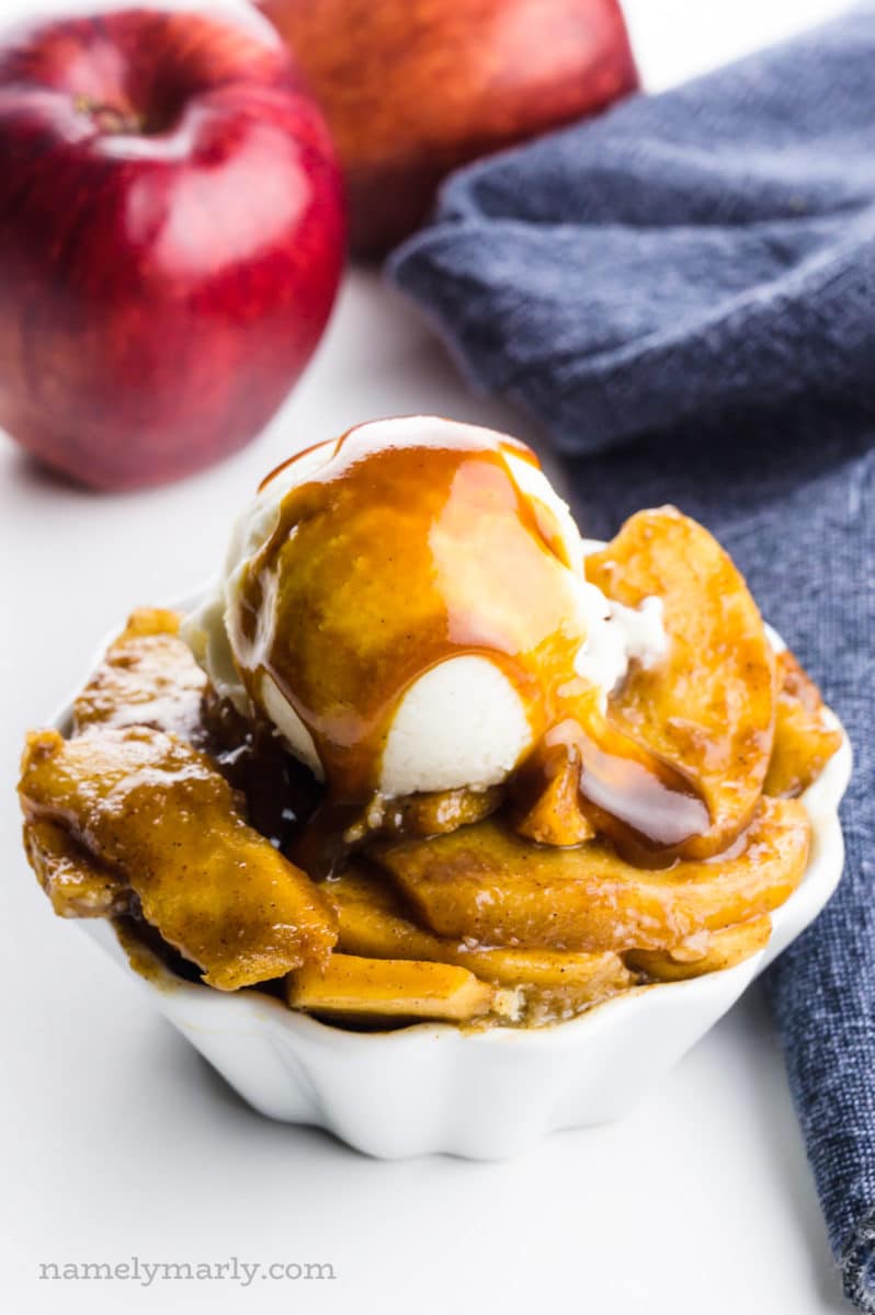 A bowl of baked apple slices is topped with ice cream and caramel sauce. There are apples behind it and a blue kitchen towel.
