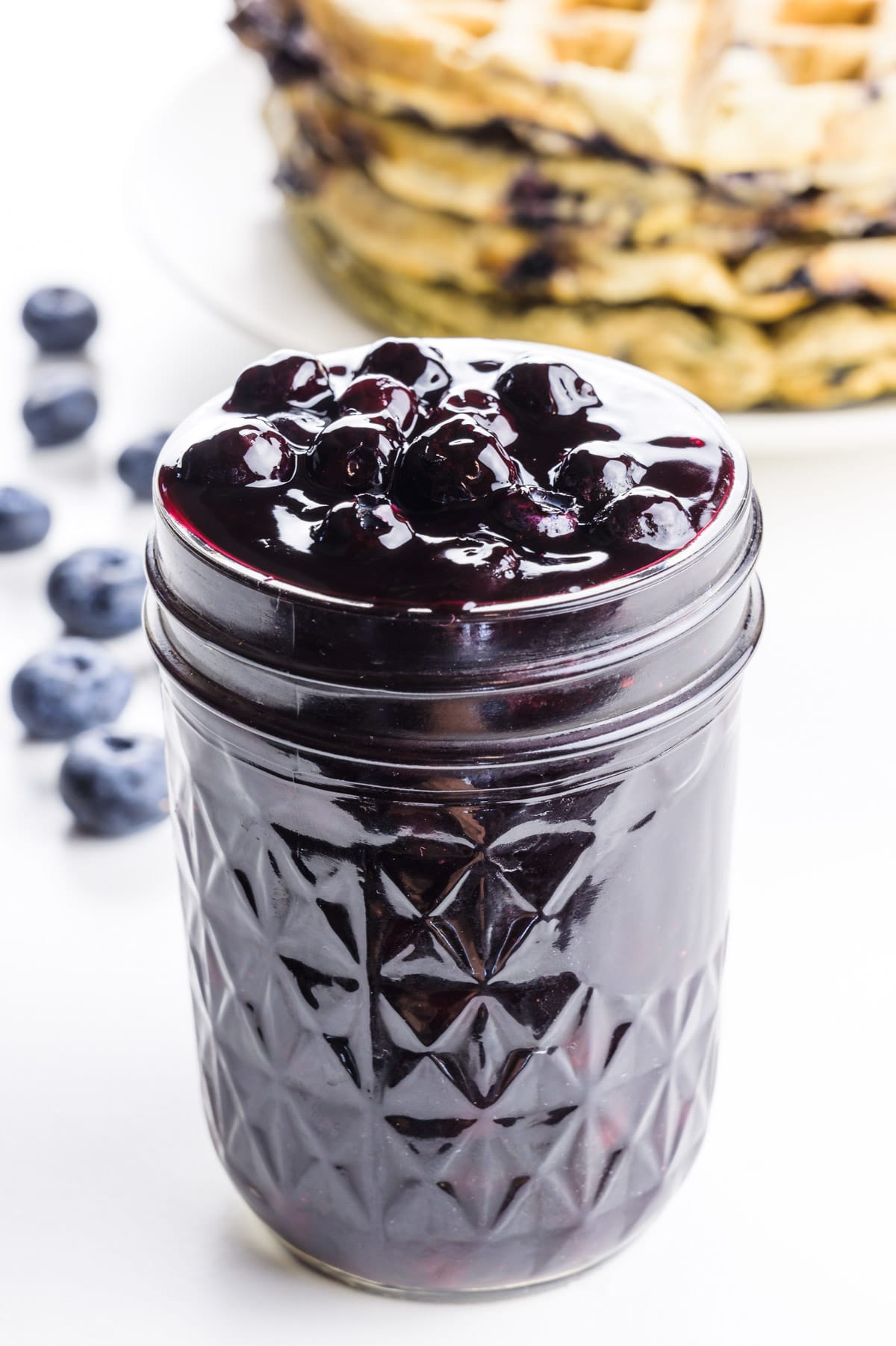 A mason jar holds blueberry sauce with blueberries and waffles behind it.