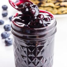 A spoon homers over a mason jar full of blueberry sauce. There are several waffles in the background.