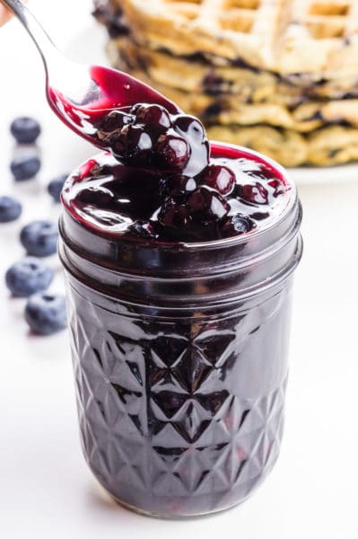 A spoon homers over a mason jar full of blueberry sauce. There are several waffles in the background.