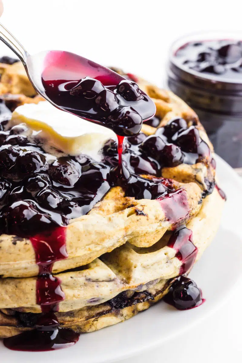 A spoon is drizzling blueberry sauce over waffles. There is a jar with more blueberry sauce behind it.