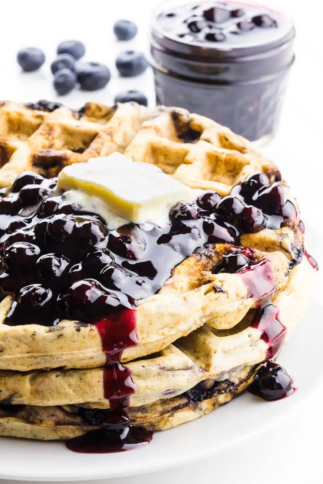 A stack of vegan blueberry waffles has vegan butter on top and some blueberry sauce. There are fresh blueberries and a bowl of more blueberry sauce behind it.