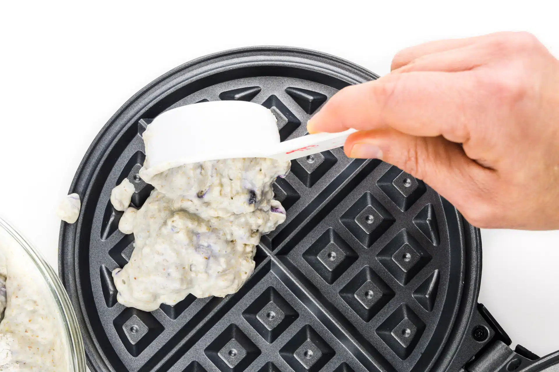 A hand holds a measuring cup pouring batter into a waffle iron.